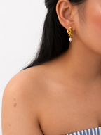 Woman wearing a sculptural gold cross stud earring with an oval grey freshwater pearl drop.