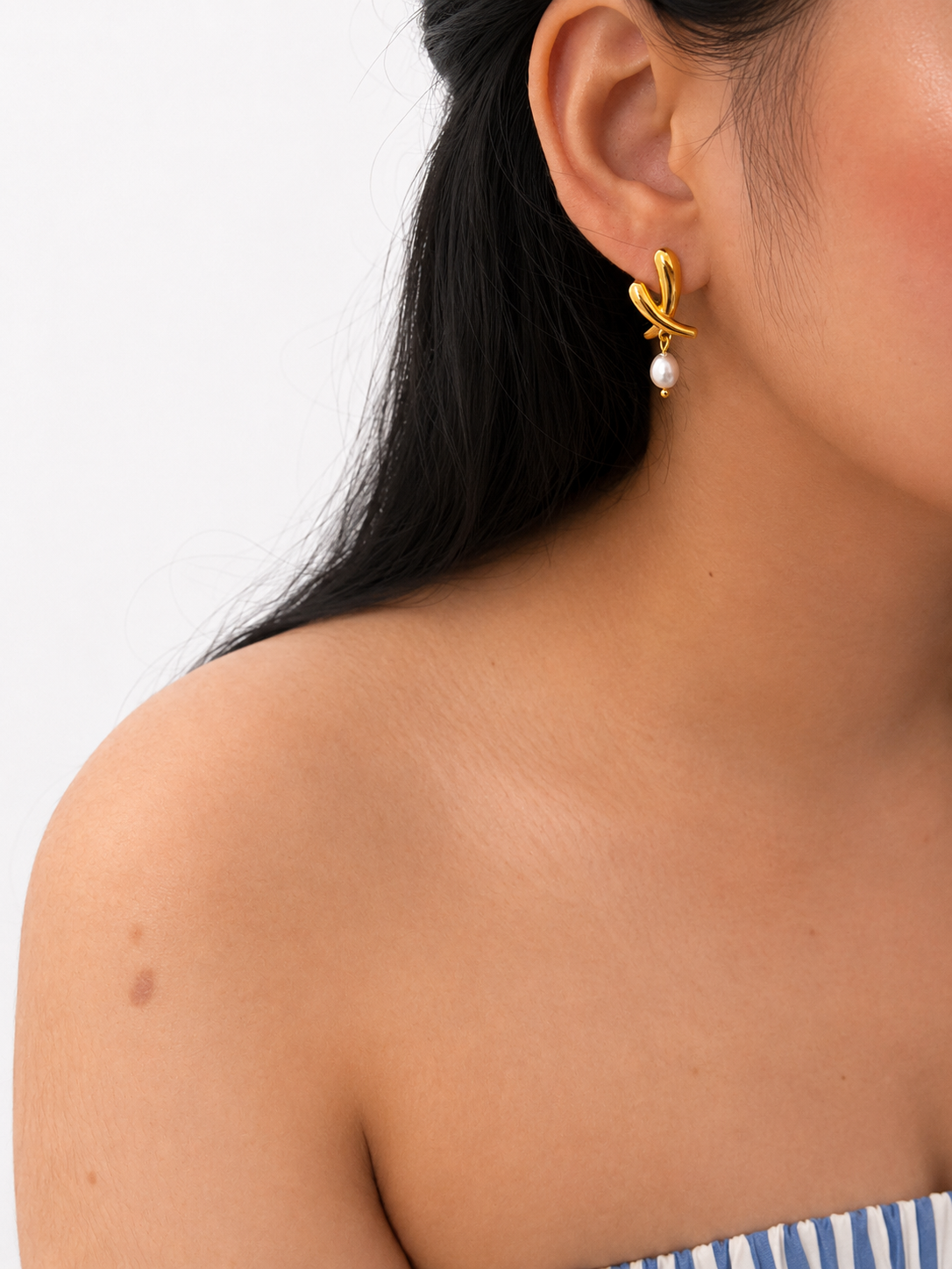 Woman wearing a sculptural gold cross stud earring with an oval grey freshwater pearl drop.