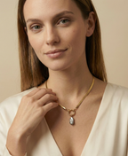 Close-up of a model wearing a flat gold herringbone chain necklace featuring a front toggle clasp and an elongated grey keshi pearl pendant, styled with a deep V-neck ivory silk top