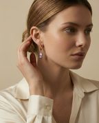 Close-up commercial jewelry photography of a gold huggie hoop earring featuring a lustrous grey keshi pearl drop. The organic, baroque-shaped pearl hangs from a sleek gold ring on a model’s ear, framed by soft blonde-brown hair. The image showcases high-quality nacre iridescence, realistic skin texture, and a warm, matte ivory background, highlighting the minimalist luxury aesthetic
