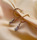 Close-up commercial jewelry photography of a gold huggie hoop earring featuring a lustrous grey keshi pearl drop. The organic, baroque-shaped pearl has high-quality nacre iridescence, realistic skin texture, and a warm, matte ivory background, highlighting the minimalist luxury aesthetic