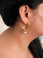 Side view of a sculptural gold hoop earring with a grey freshwater pearl drop worn on the ear.
