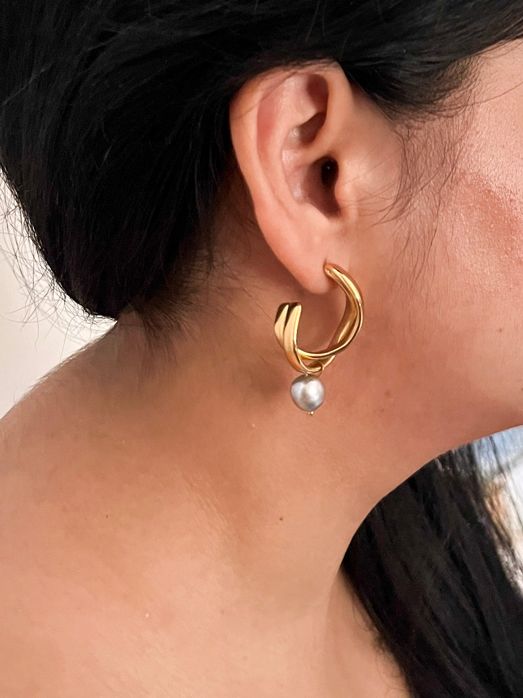 Side view of a sculptural gold hoop earring with a grey freshwater pearl drop worn on the ear.