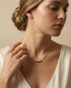 Close-up editorial portrait of a French model wearing a delicate gold chain necklace featuring three small, oval pink pearls with a high silvery luster, styled with a deep-neck ivory silk top against a matte beige background
