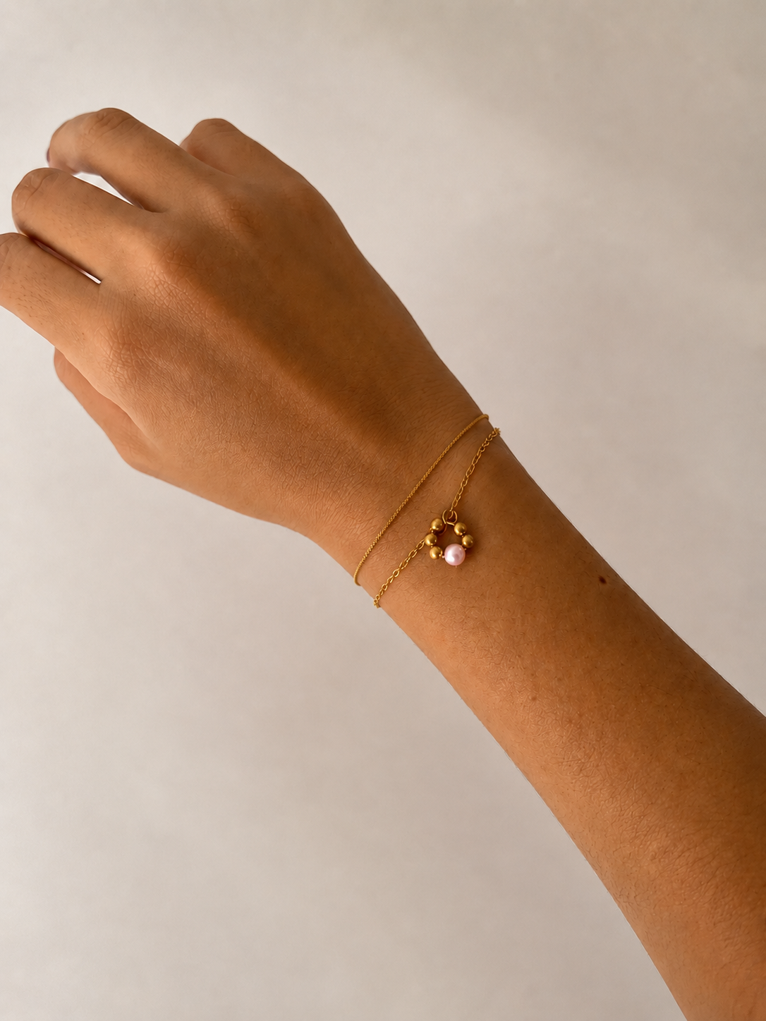 Minimalist gold double-chain bracelet with a pink freshwater pearl and circular gold bead pendant on the wrist