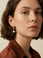Close-up of a model wearing gold drop earrings featuring a unique knot design and a dangling white round pearl. The earrings are styled with a rust-colored silk shirt.