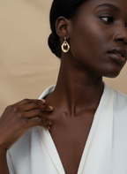 Close-up of a model wearing sculptural gold open-hoop earrings with a suspended white oval pearl. She is styled in a white silk top, showcasing elegant minimalist bridal or evening jewelry