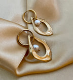 Close-up of a sculptural gold open-hoop earrings with a suspended white oval pearl. 