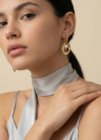 Close-up of a model wearing sculptural gold open-hoop earrings with a suspended white oval pearl. She is styled in a silver silk halter top and scarf, showcasing elegant minimalist bridal or evening jewelry.