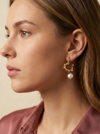 Side profile of a female model with blonde-brown hair wearing a mauve pink silk shirt and gold abstract hoop earrings featuring a round white pearl charm