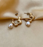 Close-up of textured gold huggie hoop earrings featuring a lustrous white pearl drop. The earrings have an organic, hammered gold finish