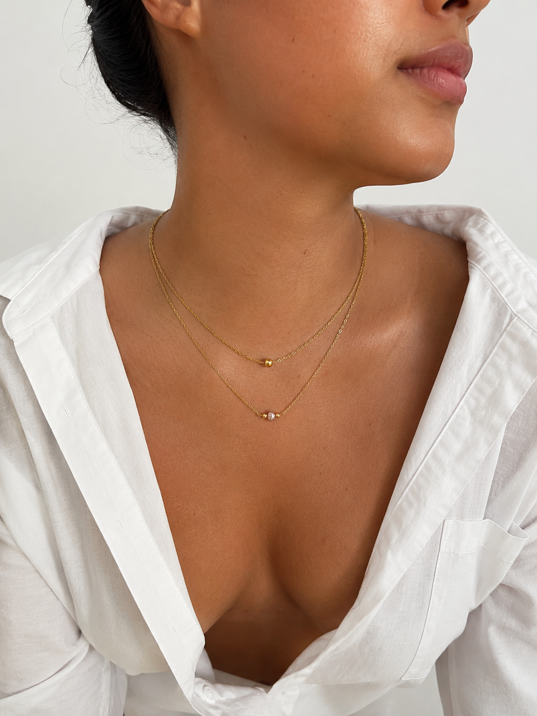 Woman wearing a minimalist gold double-layer necklace with a pink freshwater pearl and gold bead accents.