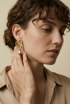 Close-up of sculptural abstract gold hoop earrings featuring a textured hammered gold leaf center and a lustrous grey pearl drop, worn by a model with curly hair for a high-end jewelry editorial