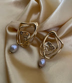 Close-up of sculptural abstract gold hoop earrings featuring a textured hammered gold leaf center and a lustrous grey pearl drop, worn by a model with curly hair for a high-end jewelry editorial