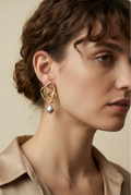 Close-up luxury jewelry editorial featuring sculptural gold open-frame earrings with a lustrous grey pearl drop. The model, with blonde curly hair, wears an ivory silk scarf and camisole, highlighting the textured gold metal and silver iridescent pearl against a soft warm studio background