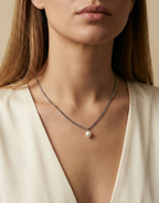 Close-up of a delicate silver curb chain necklace featuring a single white baroque pearl pendant with high luster, worn by a female model in an ivory silk top, showcasing the necklace length and elegant draping