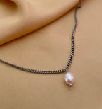 Close-up of a delicate silver curb chain necklace featuring a single white baroque pearl pendant with high luster, worn by a female model in an ivory silk top, showcasing the necklace length and elegant draping