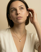 Close-up detail of a dainty gold chain necklace featuring small beads and a single lustrous white baroque pearl pendant, worn by a model in a v-neck ivory silk blouse