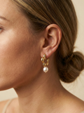 Close-up of textured gold huggie hoop earrings featuring a lustrous white pearl drop. The earrings have an organic, hammered gold finish and are worn by a model with a silver silk scarf, demonstrating the scale and elegant drape