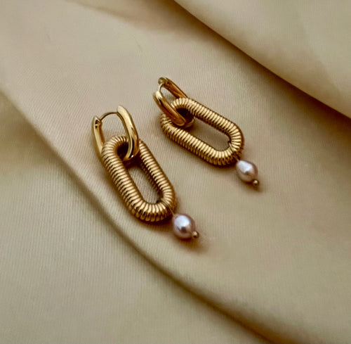 Close-up of textured gold oval link earrings featuring a dainty pink pearl drop. High-resolution commercial jewelry photography showcasing ribbed gold detailing and lustrous pearl against a soft matte ivory background.