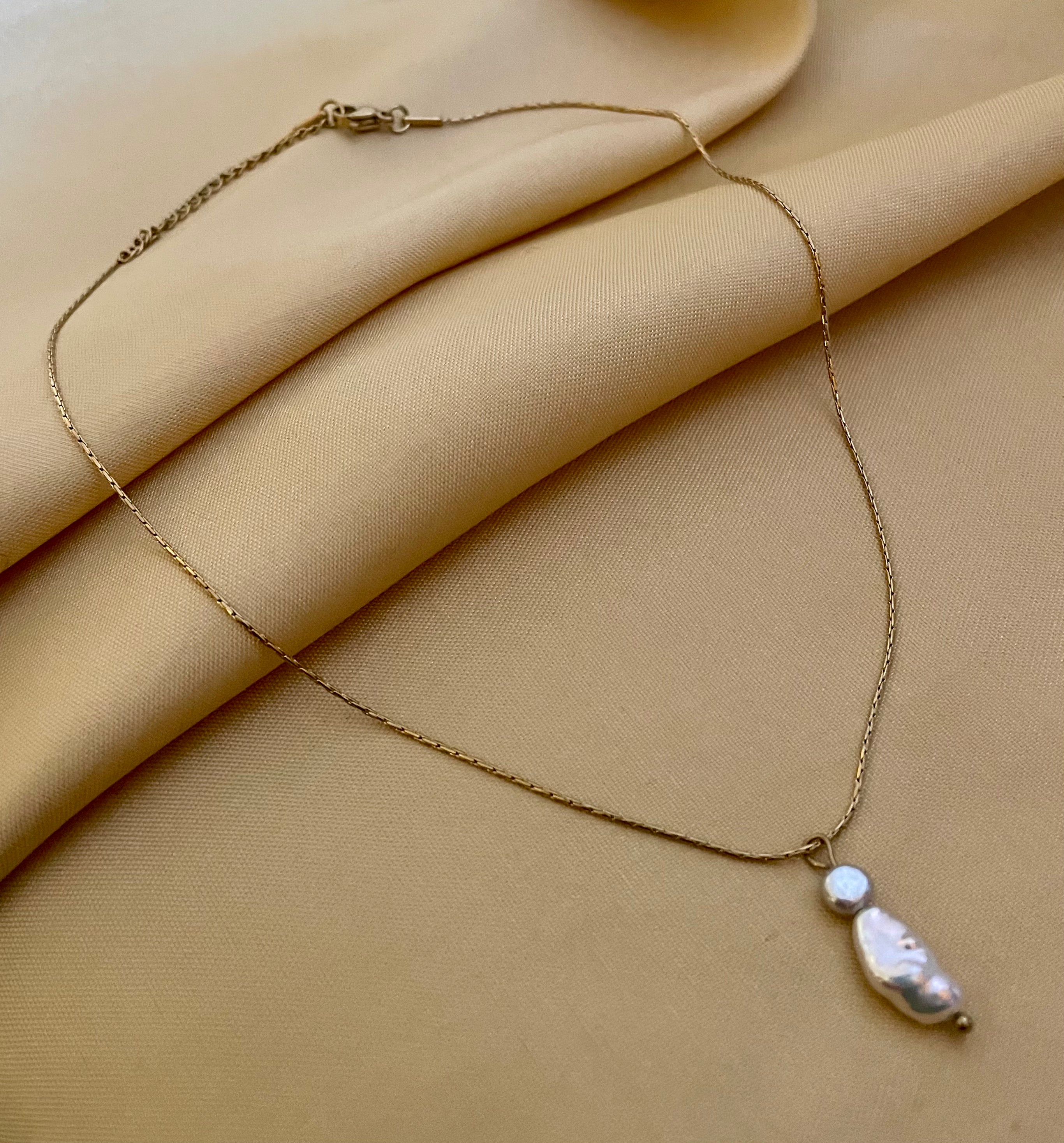 Close-up editorial portrait of a woman wearing a delicate gold chain necklace featuring a stacked pendant with a lustrous white keshi pearl and a small grey baroque pearl, styled with an ivory silk top.