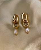Close-up of sculptural gold link earrings featuring a twisted rope texture and hanging freshwater baroque pearl drops. The earrings are displayed on a champagne silk fabric to highlight the pearl luster and high-shine gold finish