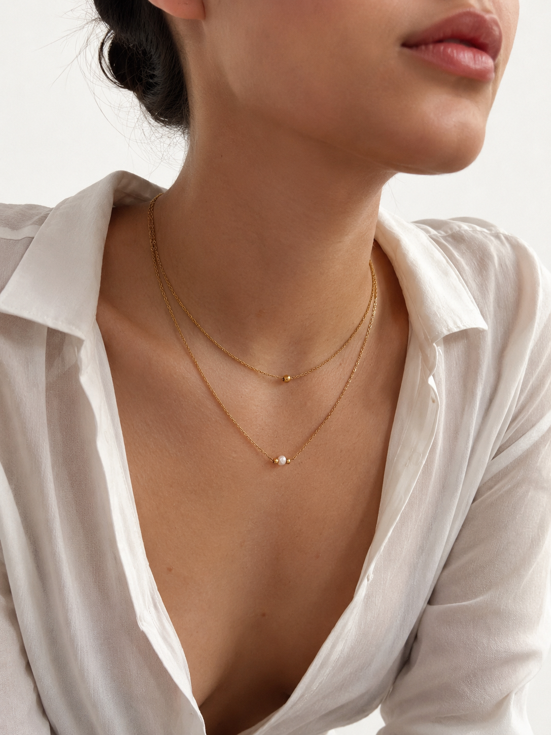 Woman wearing a minimalist gold double-layer necklace with a white baroque freshwater pearl and gold bead accents.