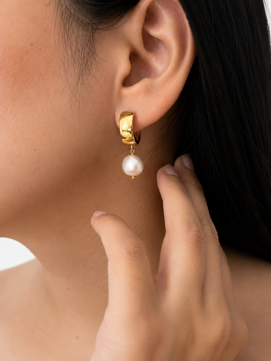 Close-up of a gold huggie earring with a white baroque freshwater pearl drop worn on the ear.