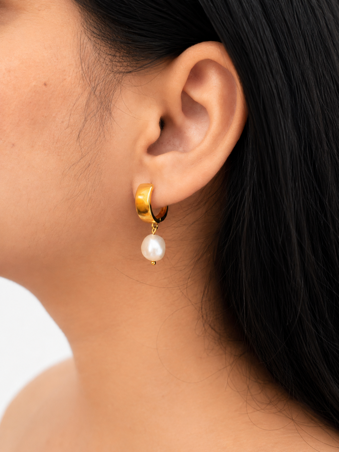 Woman wearing a polished gold huggie hoop earring with a white baroque freshwater pearl drop