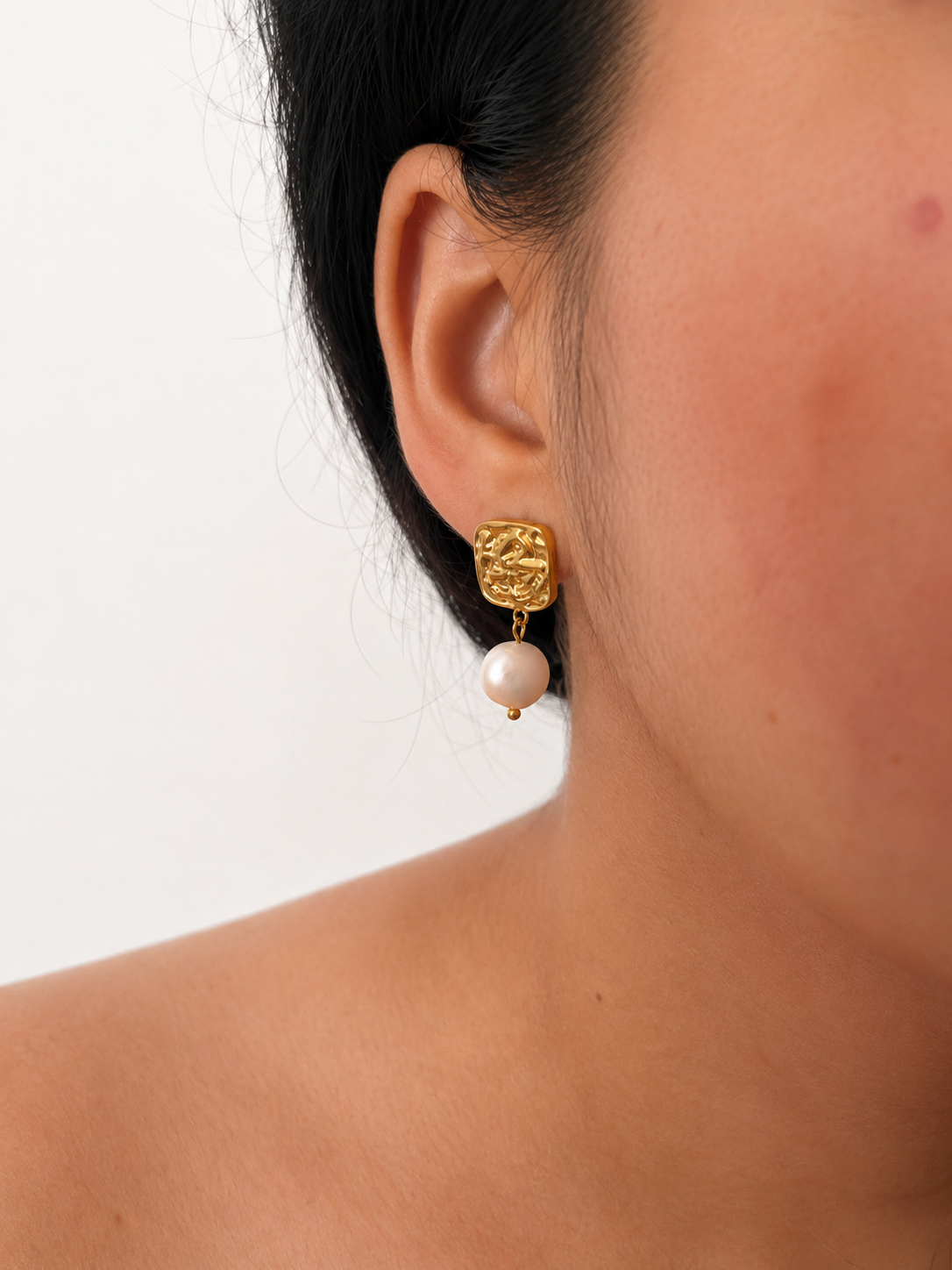 Close-up of a textured gold square earring with a white baroque freshwater pearl worn on the ear