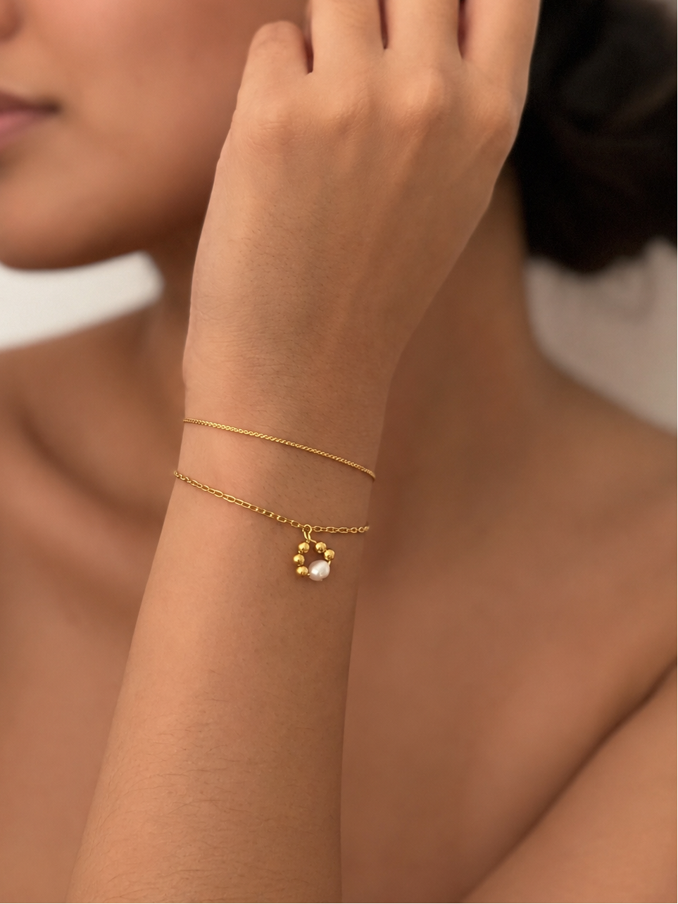 Woman wearing a gold double-chain bracelet with a petite white freshwater pearl pendant and polished gold beads