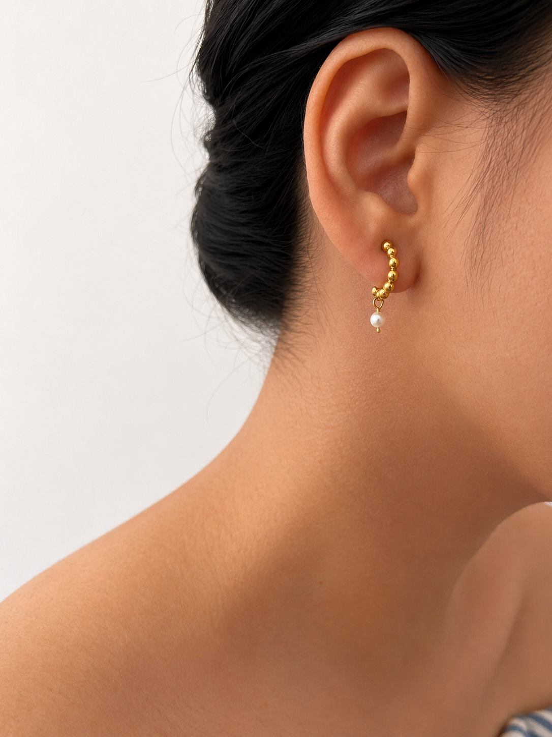Close-up of a beaded gold huggie earring with an oval white freshwater pearl worn on the ear