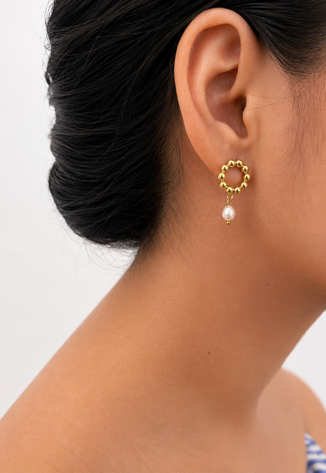 Close-up of a beaded gold circle stud earring with a white freshwater pearl drop worn on the ear.