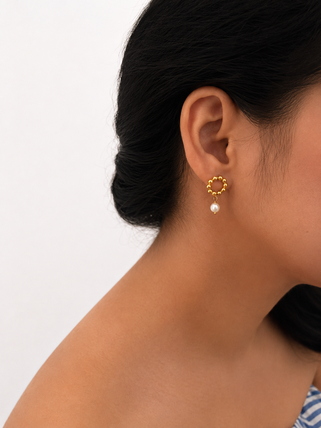Woman wearing a beaded gold circle stud earring with a white freshwater pearl drop.
