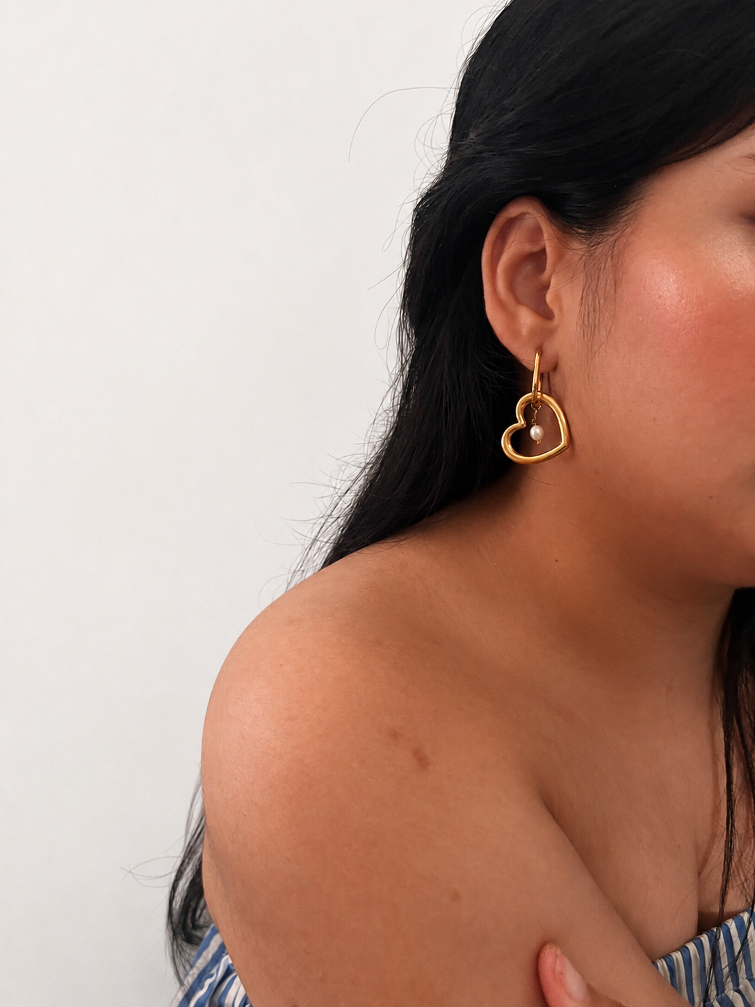 Woman wearing a gold interlinked heart earring with an oval white freshwater pearl drop.