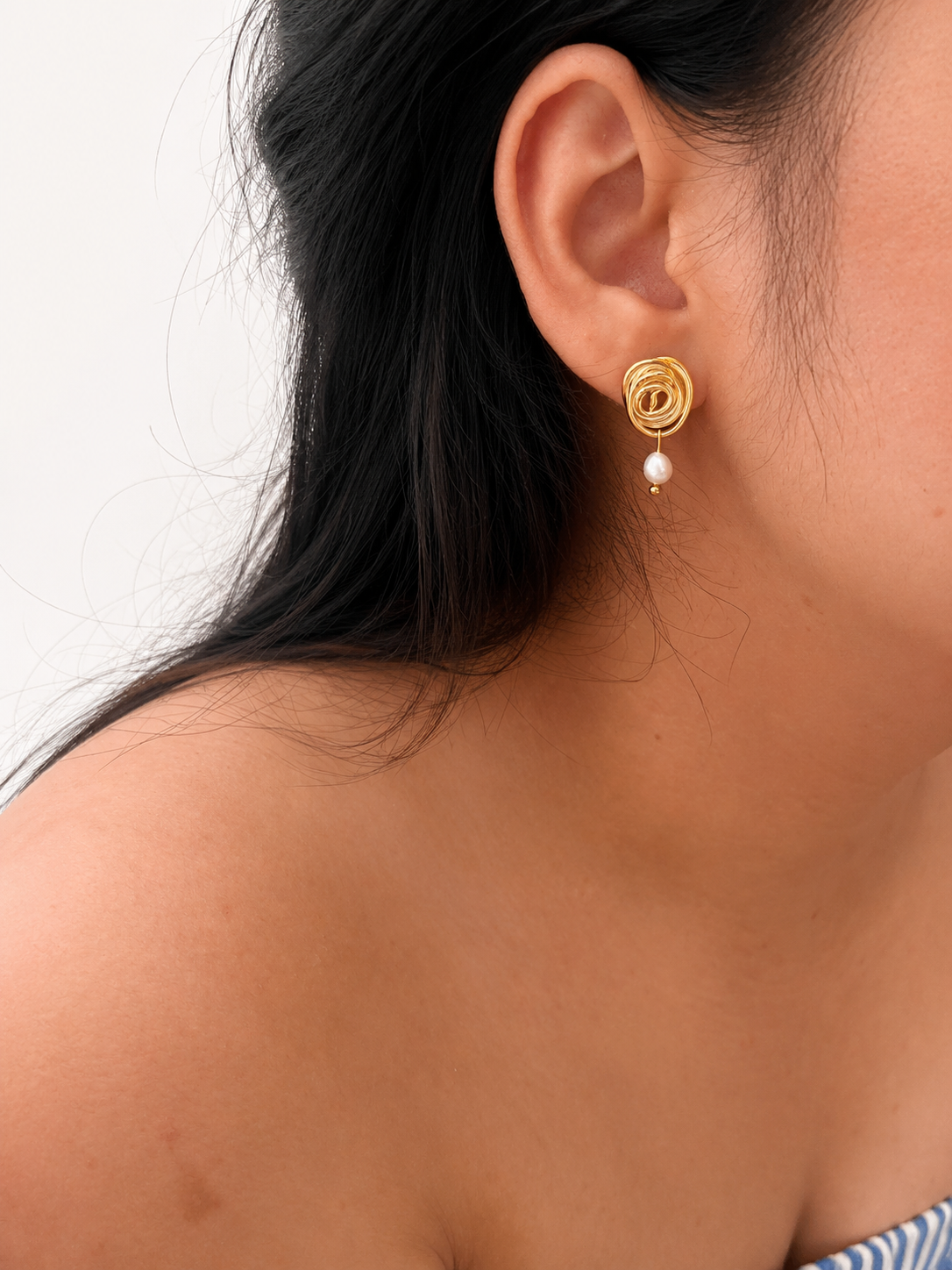 Close-up of a gold spiral stud earring with an oval white freshwater pearl drop worn on the ear