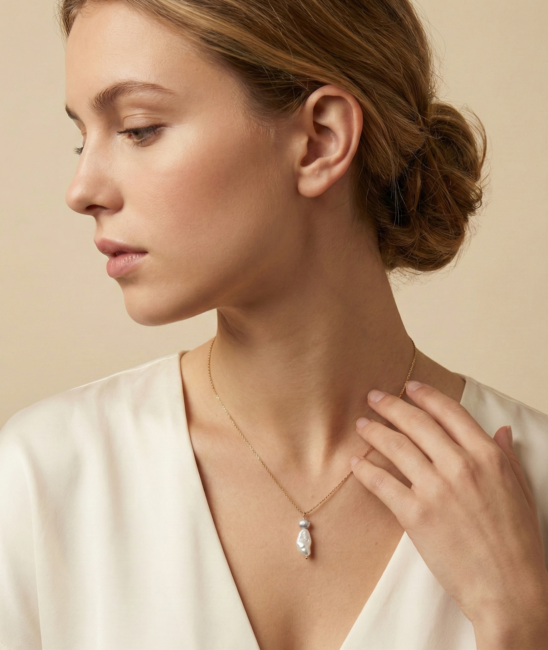 Close-up editorial portrait of a woman wearing a delicate gold chain necklace featuring a stacked pendant with a lustrous white keshi pearl and a small grey baroque pearl, styled with an ivory silk top.